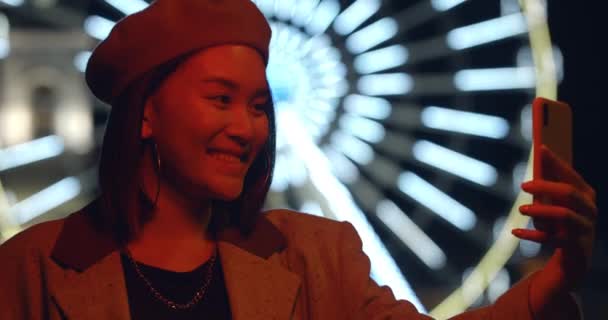 美しい若い女性が夜道で自撮りやSNSに投稿しています。ベレー帽の笑顔とスマートフォンを使用して100万人の女性。背景にライト付き観覧車. — ストック動画