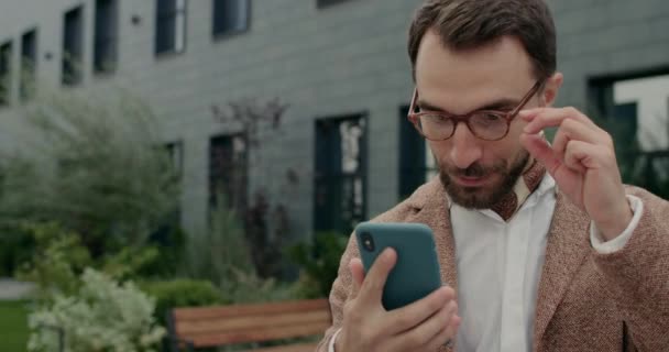 Detailní záběr šťastný muž v brýlích kontrolu obsahu smartphone a slaví úspěch. Stylový vousatý chlap s krkem, takže vzrušený výraz obličeje a křik při použití telefonu. — Stock video