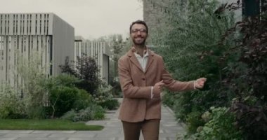 Gözlüklü ve fularlı neşeli bir adamın şehir caddesinde yürürken el çırpması ve dans etmesi. Havalı sakallı adam heyecanlı görünüyor ve gülümsüyor..