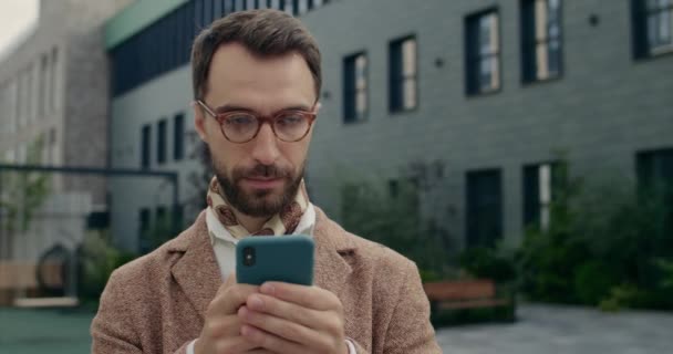 Närbild av seriös man i snygg kostym med smartphone och titta åt sidan. Snygg självsäker kille i glasögon tänker när du skriver meddelande på telefonskärmen och står på stadens gata. — Stockvideo