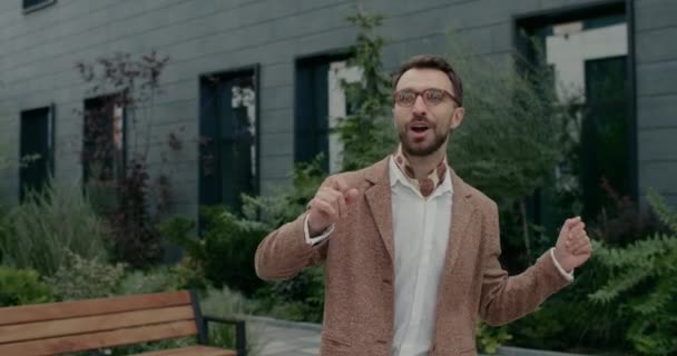 Happy guy in glasses and neckerchief singing and dancing while walking at city street. Stylish bearded male person adjusting suit and smiling while having good mood outdoors. — Vídeo de stock