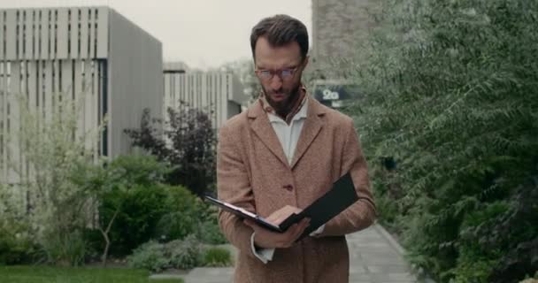 Ongelukkige man met een baard in een bril die een map vasthoudt terwijl hij financiële documenten leest. Stijlvolle zakenman kijkt teleurgesteld tijdens het lopen en het controleren van de winst in de stad straat. Begrip "bedrijf". — Stockvideo