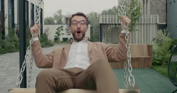 Crop view of bearded guy in glasses swinging and laughing while looking to camera. Man with neckerchief having fun while riding at swing set at near office buildings. Concept of emotions. — Vídeos de Stock