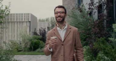Deri kağıt klasör ve kahve fincanı taşıyan gözlüklü yakışıklı bir adam. Eşarplı neşeli sakallı adam yan bakıyor ve sokakta yürürken gülümsüyor..