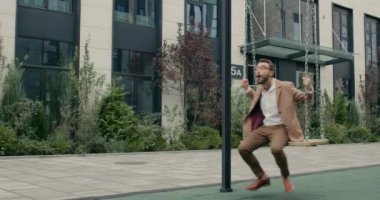 Gözlüklü yakışıklı sakallı adam sallanıyor ve gülüyor. Ofis binalarının yanındaki salıncakta sallanırken atkıyla eğlenen şık, güler yüzlü bir işadamı. Duyguların kavramı.