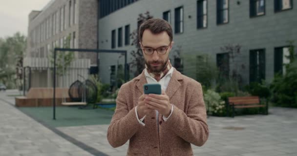Προβολή των καλλιεργειών γενειοφόρος τύπος σε κομψό κοστούμι χρησιμοποιώντας το smartphone και κοιτάζοντας στην άκρη. Όμορφος επιχειρηματίας με γυαλιά σκέψης κατά την πληκτρολόγηση μήνυμα στην οθόνη του τηλεφώνου και στέκεται στο δρόμο της πόλης. — Αρχείο Βίντεο