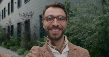 Kameraya bakarken gözlüklü, el sallayan ve konuşan neşeli adama yaklaş. Sakallı adamın gülümseyen ve barış işareti gösteren bir kamera görüntüsü..