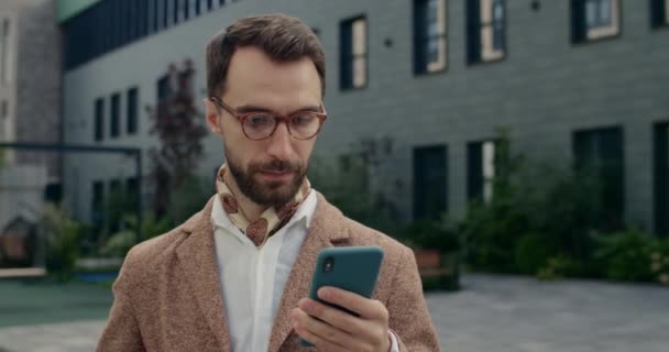 Zamknij widok pewny siebie biznesmen w stylowym garniturze za pomocą smartfona. Przystojny, poważny facet w okularach przewijający ekran telefonu, stojący na ulicy. Koncepcja sukcesu. — Wideo stockowe