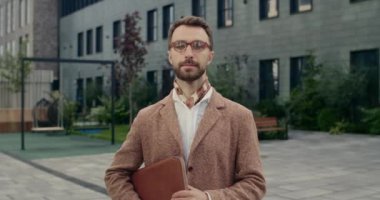 Gözlüklü yakışıklı iş adamının portresi başını kaldırıyor ve kameraya bakıyor. Sokakta dikilirken boynunda deri klasör olan şık sakallı bir adam. Açık havada..