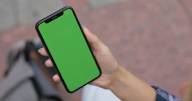 Sokakta otururken telefonlu bir kadının en iyi görüntüsü. Dikey olarak yeşil şablon ekranlı akıllı telefonu tutan kadın. Krom anahtar kavramı, taklit et. Açık havada..