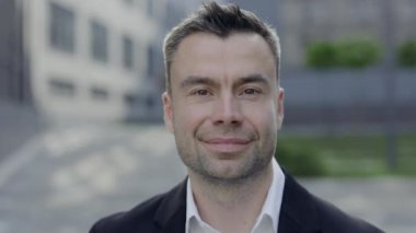 Olgun girişimcinin kameraya bakıp gülümsemesine yakından bakın. 40 'lı yaşlarda takım elbiseli yakışıklı bir adam ofis binalarının yanında poz veriyor. Başarı kavramı.