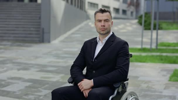 Ritratto di uomo serio in abito formale che guarda alla macchina fotografica mentre si siede sulla sedia a rotelle. Vista Crop di bello uomo d'affari disabili in posa vicino edificio per uffici. — Video Stock