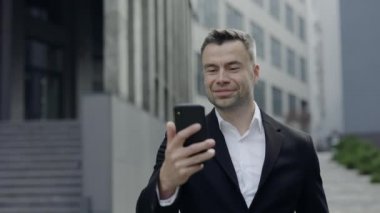 Klasik takım elbiseli, neşeli ve akıllı telefon ekranına bakan mutlu olgun bir adamın görüntüsü. 40 'lı yaşlardaki yakışıklı işadamı sokakta yürürken başarıyı kutluyor ve gülümsüyor..