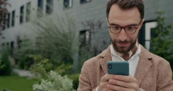 Close-up van de man in glazen controleren smartphone-inhoud en verheugen. baard man met halsdoek het maken van opgewonden gezichtsuitdrukking tijdens het gebruik van telefoon en lopen op straat. — Stockvideo