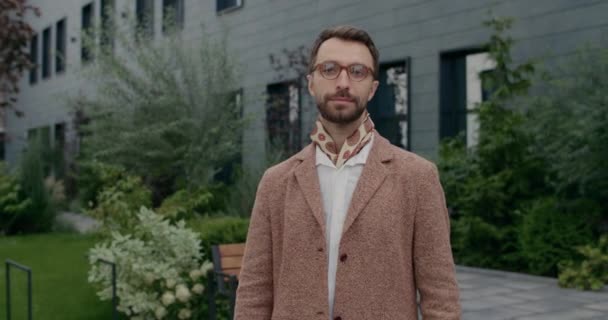 Portrait of cheerful guy in glasses and neckerchief turning head and crossing arms. Handsome bearded businessman in stylish clothes posing and smiling while standing at city street. — Stock Video