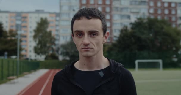 Vista de la cosecha de joven hombre serio girando la cabeza y mirando a la cámara. Retrato de un hombre reflexivo de pie en el estadio de deportes al aire libre. Concepto de deporte, motivación. — Vídeos de Stock