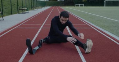 Spor sahasında otururken protez koşu bıçaklarıyla gerinen sakat erkek sporcu. Ampüte edilmiş bacağı ısınıyor ve öne eğiliyor. Özürlü kavramı, motovasyon.