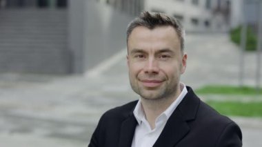 Olgun iş adamının kameraya bakışını yakından izle. 40 'lı yaşlarda takım elbiseli, açık havada ofis binalarının yakınında gülümseyen neşeli bir adamın portresi. İş kavramı.