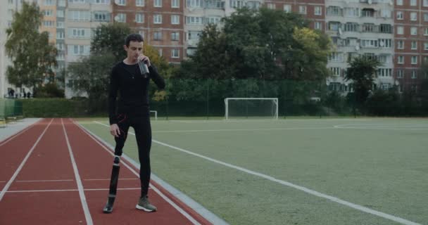 Αναπήρων νεαρός με προσθετικές λεπίδες πίνοντας νερό από αθλητικό μπουκάλι. Άφθονος αρσενικός δρομέας στέκεται στο γήπεδο και κοιτάζει μπροστά. Έννοια του κινητικού αθλητικού υλικού. — Αρχείο Βίντεο