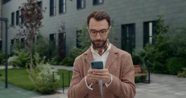 Bonito cara de óculos olhando para o lado enquanto digita na tela do smartphone. Vista de corte de homem barbudo elegante com lenço usando telefone para navegar na internet enquanto caminha na rua. — Vídeo de Stock