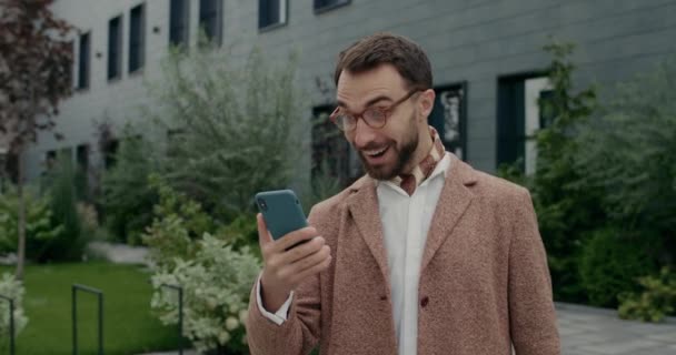 Vzrušený muž v brýlích má dobré zprávy na svém smartphonu, zatímco se raduje a dělá ano gesto. Stylový vousatý podnikatel s oslavným úspěchem a úsměvem při použití telefonu. — Stock video