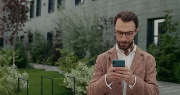 Chlápek v brýlích se dotýká obrazovky při kontrole obsahu smartphonu. Vousatý muž s krkem radovat a dělat překvapený výraz obličeje při použití telefonu a chůze na ulici. — Stock video