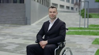 Tekerlekli sandalyede otururken kameraya bakan resmi takım elbiseli neşeli bir adamın portresi. Ofis binası yakınlarında kameraya poz veren yakışıklı engelli yönetim kurulu başkanı görüntüsü.