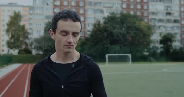 Vista de la cosecha de hombre joven serio beber agua de la botella y mirando hacia el futuro. Retrato de un hombre reflexivo de pie en el estadio de deportes al aire libre. Concepto de deporte, motivación. — Vídeos de Stock