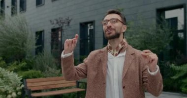 Şehir caddesinde yürürken şarkı söyleyen ve dans eden gözlüklü neşeli adam manzarası. Sakallı şık bir adam takım elbisesini ayarlıyor ve keyfi yerindeyken kafiyeli hareket ediyor..