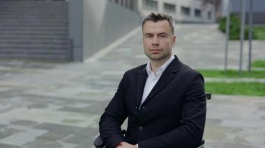 Resmi takım elbiseli ciddi bir adamın portresi başını çeviriyor ve tekerlekli sandalyede otururken kameraya bakıyor. Ofis binasının yakınındaki engelli iş adamının mahsul manzarası. Yakınlaştır.