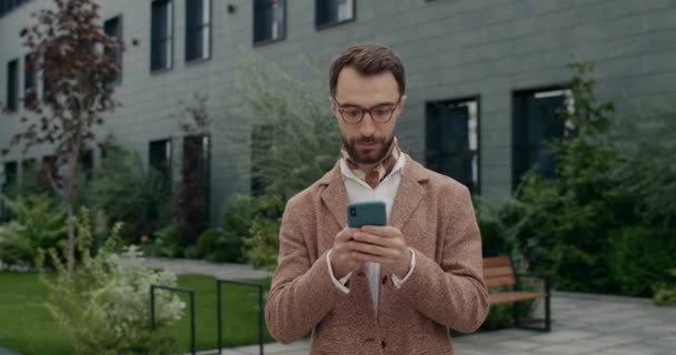 Schnitte Ansicht von ernsten Kerl in stilvoller Kleidung mit Smartphone und Blick zur Seite. Stilvoller Geschäftsmann mit Brille tippt Nachricht am Telefonbildschirm ein, während er auf der Straße spaziert. — Stockvideo