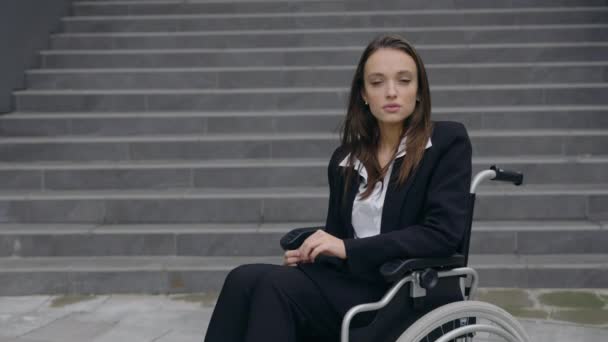 Портрет інвалідної серйозної бізнес-леді в костюмі, сидячи в інвалідному візку на вулиці. Інвалідність приваблива жінка повертає голову і дивиться на камеру на відкритому повітрі. Збільшити масштаб . — стокове відео