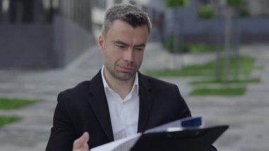 Tekerlekli sandalyedeki erkeğin şehir caddesindeki mali raporlara baktığı yakın plan görüntüsü. Takım elbiseli başarılı bir işadamı belge okuyor ve dışarıda mutlu görünüyor..
