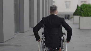 Sokakta tekerlekli sandalyede oturan takım elbiseli adamın arkadan görünüşü. Ofis binasının yakınında çalışan engelli bir erkek. Motivasyon kavramı, ızgara.