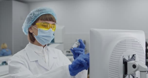 Großaufnahme einer Forscherin in Schutzmaske und Google-Brille, die Daten am Computer eingibt. Wissenschaftler in Raub und Bouffant Verschluss hält Glas medizinische Flasche während der Eingabe auf dem Bildschirm im Labor. — Stockvideo