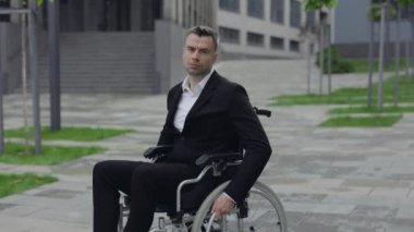 Şehir caddesinde tekerlekli sandalyede otururken klasik takım elbiseli engelli bir iş adamı. 40 'lı yaşlarda ciddi engelli bir erkek kameraya bakıyor. Açık havada..