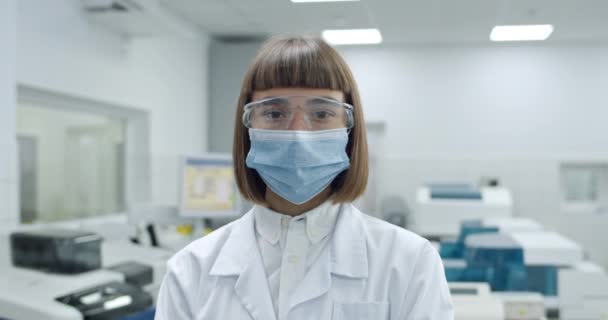 Πορτρέτο της νεαρής γυναίκας με προστατευτική μάσκα και googles κοιτάζοντας στην κάμερα. Άποψη των καλλιεργειών των γυναικών σε λευκό επαγγελματικό παλτό posingwhile στέκεται στο εργαστήριο. Έννοια της ιατρικής. — Αρχείο Βίντεο