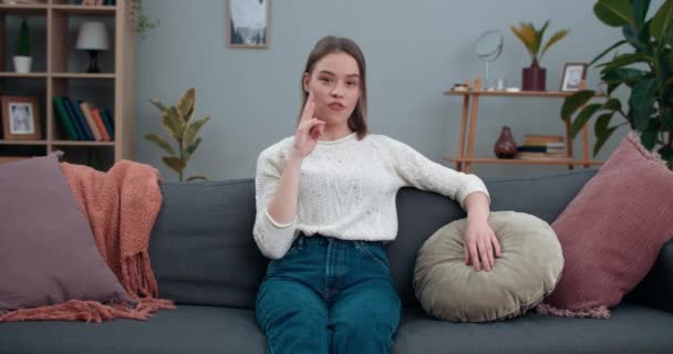 Ung vacker kvinna visar med dövstum teckenspråk blir vegan och tittar till kameran. Vacker kvinnlig person som främjar vegetarianism medan du sitter på soffan hemma. — Stockvideo