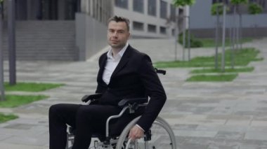 Sokakta tekerlekli sandalyede otururken klasik takım elbiseli başarılı bir iş adamı. Neşeli engelli erkek kameraya bakarken arkasını dönüyor ve gülümsüyor.