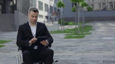 Resmi takım elbiseli bir adamın şehir caddesinde tekerlekli sandalyede otururken tablet kullanması ve sevinmesi. Engelli mutlu işadamı gülümsüyor ve ofis binasının yakınında evet hareketi yapıyor..