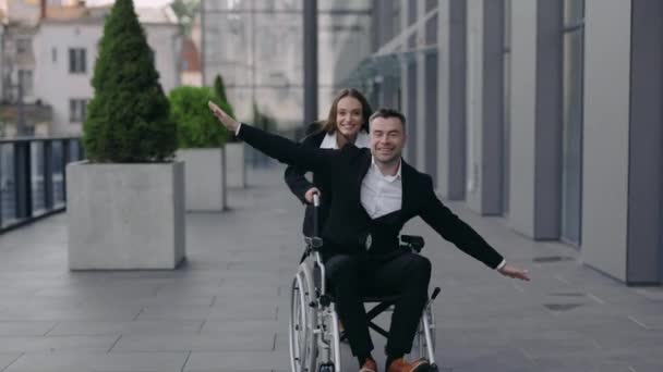 Веселая молодая женщина, толкающая инвалидное кресло со своим коллегой-инвалидом, улыбаясь возле офисного здания. Мужчина и женщина счастливые люди веселятся вместе на улице. Концепция поддержки. — стоковое видео