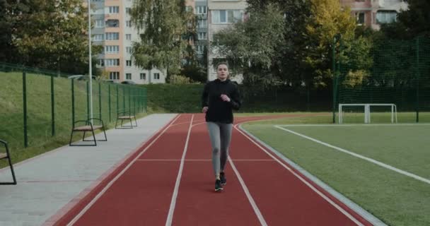 Attraktive Millennial-Frau, die nach vorne blickt und beim Joggen tief durchatmet. Hübsche Frau im Trainingsanzug läuft auf dem Sportplatz. Konzept eines gesunden Lebensstils, Motivation. — Stockvideo