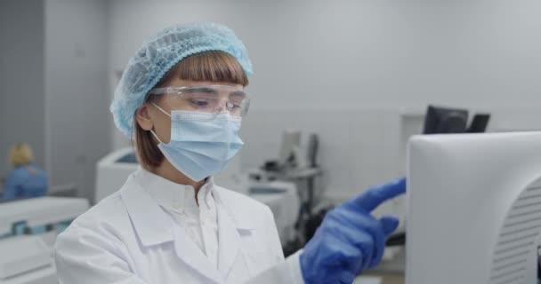 コンピュータにデータを入力する保護マスクやグーグルで女性医療従事者の閉鎖。ロブの科学者と研究室で入力しながらガラス試験サンプルチューブを保持する浮力キャップ. — ストック動画