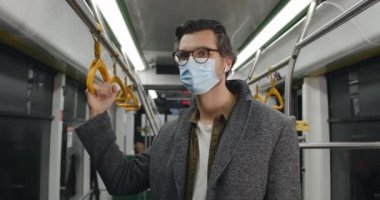 Koruyucu tıbbi maskeli bir adamın portresi başını çevirip kameraya bakıyor. Kablosuz kulaklıklı bir erkek toplu taşıma aracıyla giderken tırabzan tutuyor. Salgın kavramı.