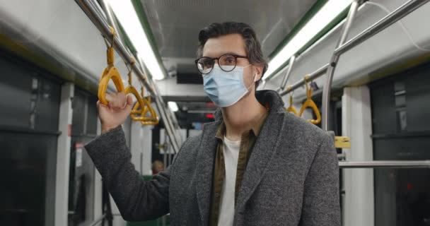 Porträtt av killen i skyddande medicinsk mask vrida huvudet och titta på kameran. Man i trådlösa hörlurar håller ledstång medan du går i kollektivtrafik. Begreppet pandemi. — Stockvideo