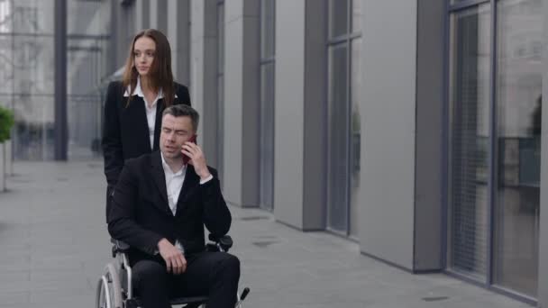 当一个女人和她的残疾老板一起推着轮椅在办公大楼附近行走时的倒影。男首席执行官和他的女秘书一起在智能手机上交谈时提供帮助. — 图库视频影像