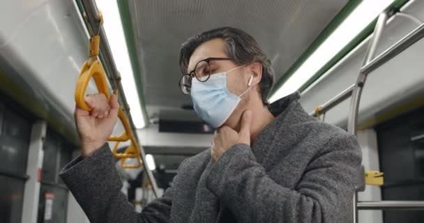 Парень в защитной медицинской маске кашляет и замерзает, стоя в подземном поезде. Мужчина, больной в беспроводных наушниках плохо себя чувствует в общественном транспорте. Понятие пандемии, болезни. — стоковое видео