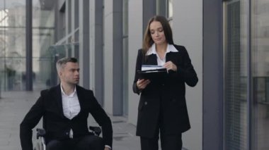 Tekerlekli sandalyedeki geçersiz erkek CEO ve ofis binasının yakınındaki belgelerle çalışan kadın çalışan. Şehir caddesinde yürürken iş adamlarının konuşma görüntüsü.