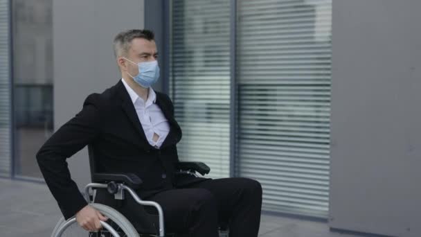 穿着防护面罩的时尚男人坐在轮椅上向前看的侧视图。男性首席执行官接近办公大楼的作物景观。大流行病的概念. — 图库视频影像