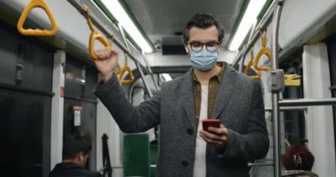 Medikal maskeli ve gözlüklü akıllı telefon kullanan yakışıklı bir adam. Kablosuz kulaklık takan yetişkin bir erkek kafa kaldırıyor ve toplu taşıma araçlarında kameraya bakıyor..